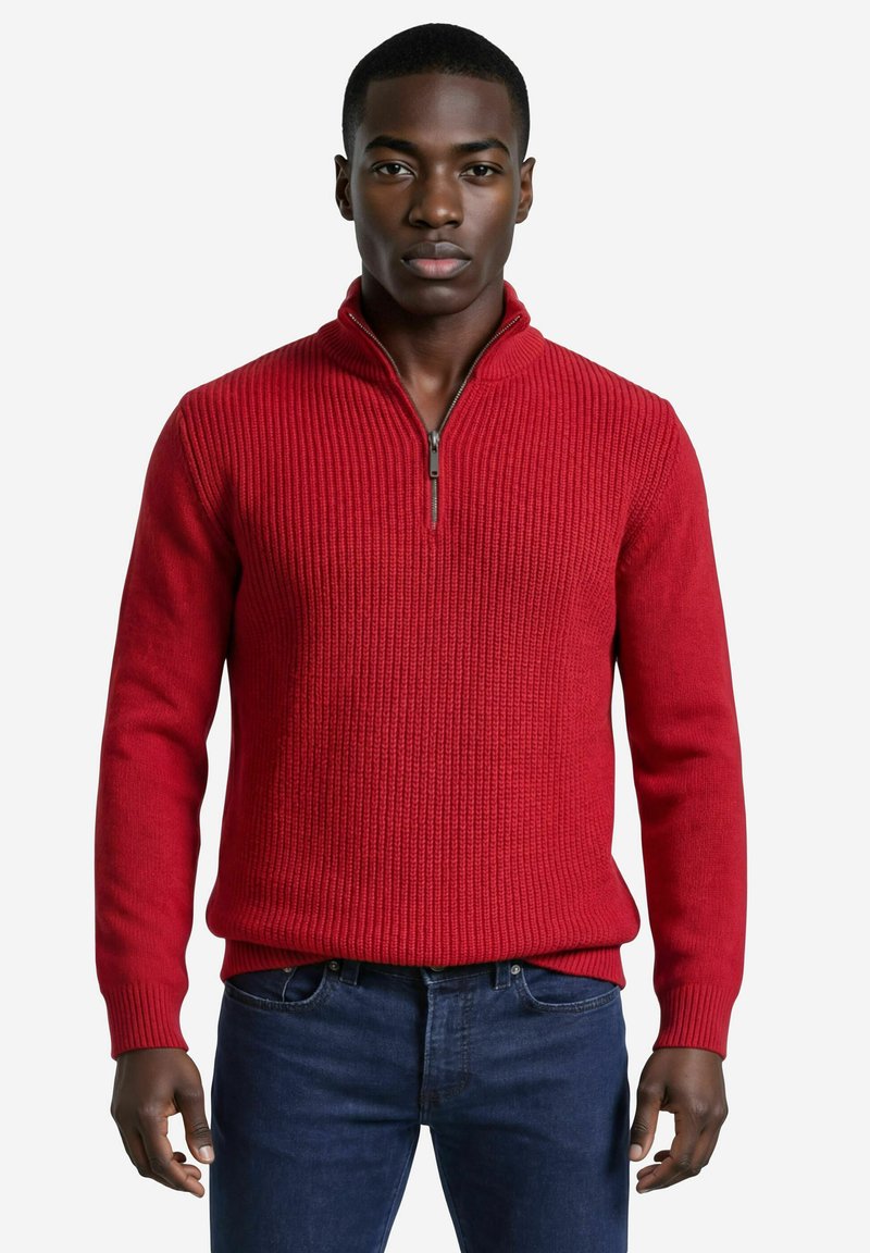 Jeune homme debout face à l'avant, portant un pull rouge côtelé à demi-fermeture éclair et un jean bleu foncé, devant un fond uni.