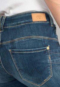 Jeans de denim azul oscuro con una vista ampliada del bolsillo trasero, que presenta una forma redondeada y sutilezas en los detalles de costura. Parche de cuero de la marca visible.