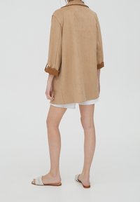 Femme portant un manteau marron clair aux manches retroussées, un short blanc et des sandales blanches à enfiler, debout devant un fond clair uni.