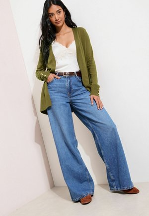 Green cardigan with button accents, white lace tank, blue wide-leg jeans, and brown suede heeled shoes against a light background.