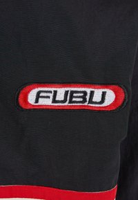 Tissu noir avec un écusson rectangulaire bordé de rouge portant les lettres FUBU en noir sur fond blanc, près d’un bord à rayures rouges et blanches.