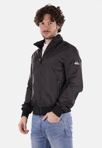 Giacca leggera nera con colletto alto, chiusura lampo frontale, tasche laterali e polsini a coste. Il materiale appare liscio e lucido.