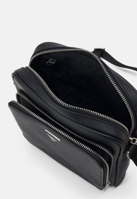 Schwarze Leder-Umhängetasche mit mehreren Reißverschlussfächern, strukturiertem Oberflächenmaterial und silbernem Logo-Akzent. Das gefütterte Innenleben weist ein sich wiederholendes Muster der Marke auf.