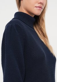Mornarsko modra teksturirana jopica s turtleneck izrezom in napihnjenimi rokavi, ki ima tkano vzorec po celotni tkanini.
