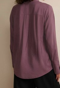 Blusa a maniche lunghe in tessuto viola tenue, dalla consistenza morbida, con orlo arrotondato e dettaglio di piega sul retro, abbinata a pantaloni neri con righe sottili.