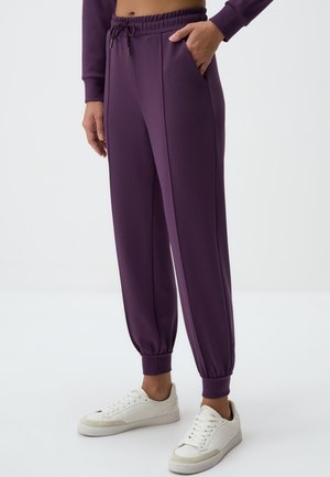 Pantaloni della tuta viola con vita e polsini elastici, dotati di due tasche laterali e una superficie liscia. Abbinati a sneakers bianche.
