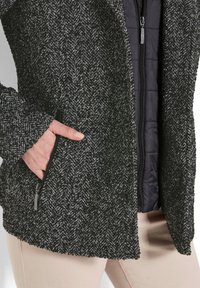 Hand in zijzak van zwart-wit geblokte ritsjack die over een zwart gewatteerd vest is gedragen, met beige broek zichtbaar aan de onderkant.