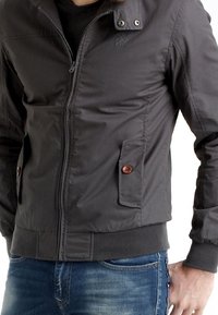 Szara kurtka zapinana na zamek z stójką, z zatrzaskami, dwoma przednimi kieszeniami z brązowymi guzikami i prążkowanym dołem. Gładka tekstura, lekki materiał.