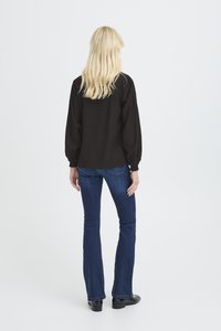 Blusa nera a maniche lunghe con polsini elasticizzati, abbinata a jeans denim blu svasati, vista da dietro. Tessuto liscio, vestibilità comoda.