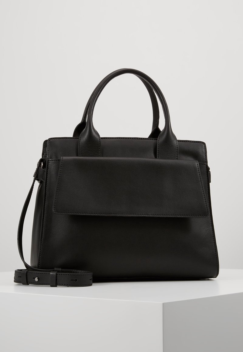 Sac à main en cuir noir avec double anses, poche avant à rabat et bandoulière amovible et réglable sur une surface blanche.