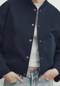 Veste courte marine en coton avec boutons en bronze, col arrondi et poches latérales, associée à un pull gris clair et un denim.