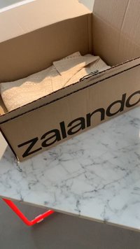 Brun papkasse fra Zalando på et marmorbord, indeholdende et lysebrunt struktureret stof. Kassen har et stort sort logo på siden.