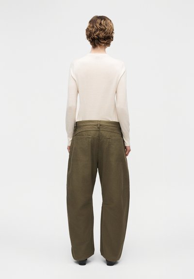 Personne aux cheveux courts et bouclés portant un haut blanc à manches longues et un pantalon large taille haute de couleur vert olive, debout de dos.