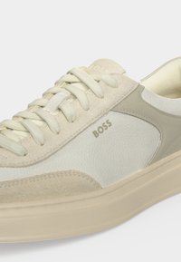 Gros plan d'une basket en cuir beige et blanc avec des détails en daim, un design à lacets et le logo "BOSS" sur le côté.