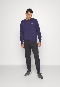 Marinblå sweatshirt med rund hals och vit logotyp, matchad med svarta joggers och sportskor med grå accenter och textilstruktur.