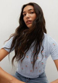 Jasnoniebieski sweter z krótkim rękawem o wzorze warkocza, ozdobiony kwiatową haftem w kolorach różowym i zielonym, zestawiony z szortami jeansowymi.