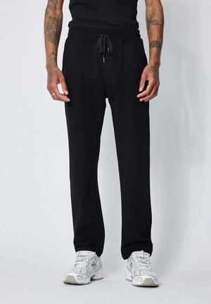 Personne portant un pantalon de jogging noir à cordon, des baskets blanches et un débardeur noir, bras le long du corps, tatouages visibles sur les deux bras.
