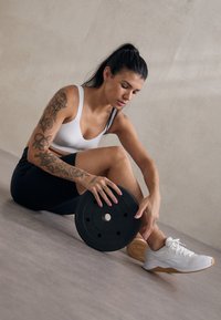 Γυναίκα με τατουάζ που κάθεται στο πάτωμα, κρατώντας μια μαύρη πλάκα βάρους, φορώντας άσπρο αθλητικό σουτιέν, μαύρες βερμούδες και άσπρα αθλητικά παπούτσια.