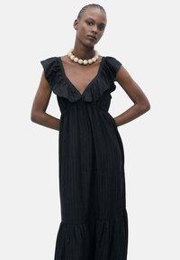 Robe maxi noire à côtes avec un décolleté en V profond froncé et des manches courtes, présentant un ourlet à volants. Accentuée par un collier en perles chunky.