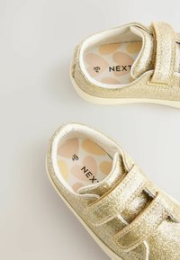 Baskets à paillettes dorées avec une sangle velcro, doublure intérieure blanche et semelle intérieure à motifs avec des designs circulaires dans des couleurs douce.