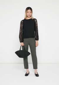Svart teksturert bluse med blondeslirer, olivengrønne, tettsittende bukser, svarte flate sko og en svart strukturert håndveske. Enkel og moderne design.