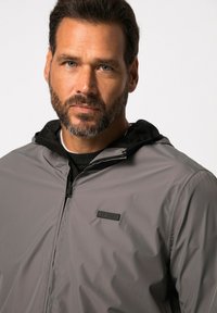 Grå zip-up jakke med hætte, lavet af let materiale. Har en sort indvendig foring og et lille præget logo på brystet.