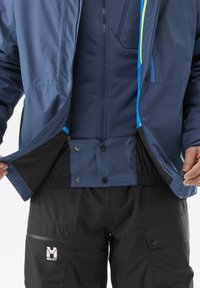 Man wearing navy blue and black outdoor jacket and black Millet brand pants, holding jacket open to show inner wind skirt with snap buttons.