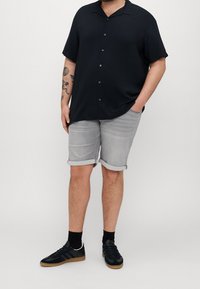 Persona vistiendo una camisa negra de manga corta con botones, pantalones cortos de mezclilla claros y enrollados, calcetas negras y zapatillas negras con suelas de goma.