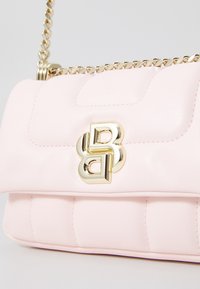 Różowa skórzana torebka z pikowanym wzorem, złotym łańcuszkiem oraz złotym akcentem logo z przodu. Gładka tekstura z zaokrąglonymi krawędziami.