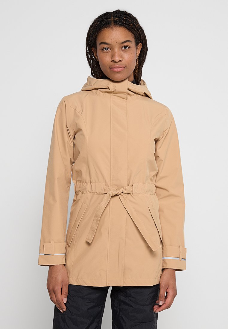 Młoda kobieta w beżowej kurtce z kapturem z marszczeniem w talii i czarnych spodniach, stojąca na tle jednolitego jasnoszarego tła.