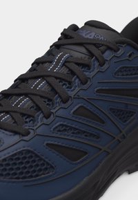 Αθλητικό παπούτσι σε ναυτικό μπλε και μαύρο με διάτρητα πάνελ, λεπτομερείς επικαλύψεις, μαύρα κορδόνια και μαλακή σόλα.