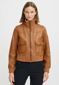 Donna che indossa una giacca di pelle beige con zip frontale e due tasche sul petto con bottone a pressione, abbinata a pantaloni neri, in piedi contro uno sfondo bianco.