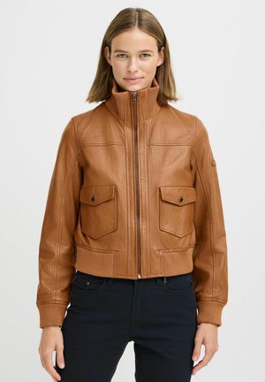 Donna che indossa una giacca di pelle beige con zip frontale e due tasche sul petto con bottone a pressione, abbinata a pantaloni neri, in piedi contro uno sfondo bianco.