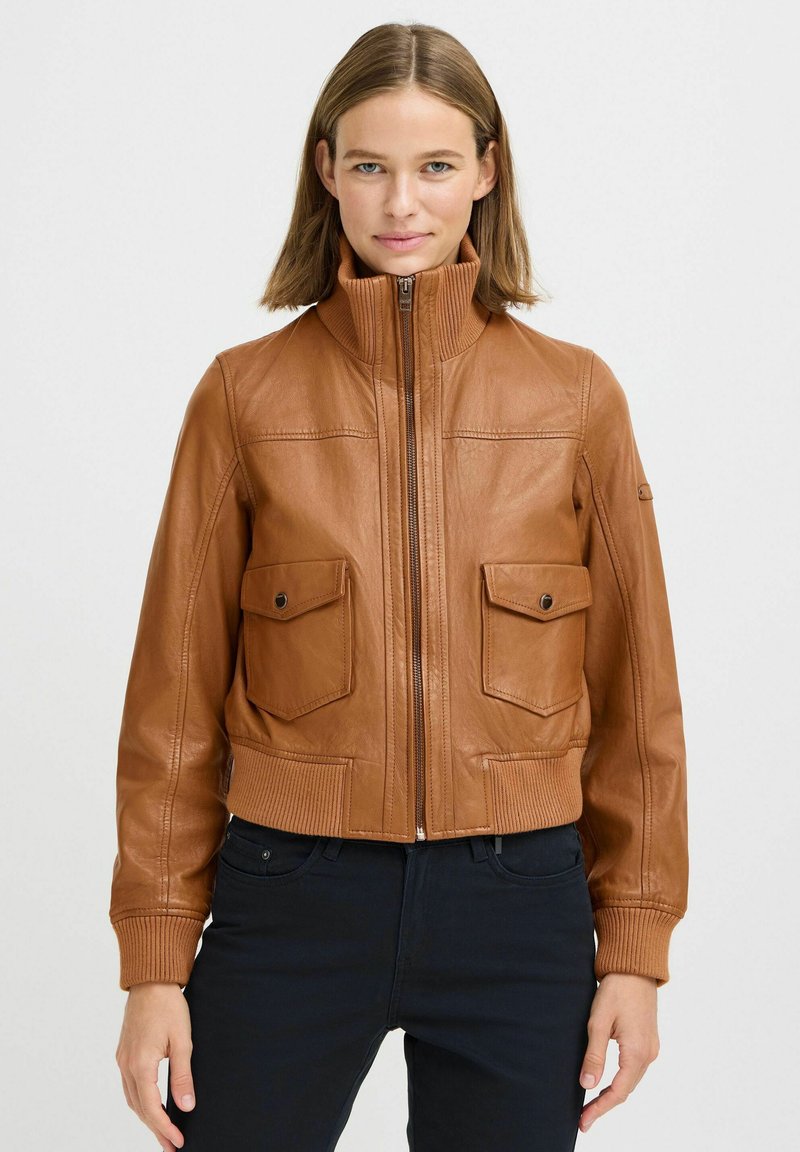Donna che indossa una giacca di pelle beige con zip frontale e due tasche sul petto con bottone a pressione, abbinata a pantaloni neri, in piedi contro uno sfondo bianco.