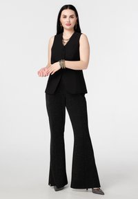 Zwarte mouwloze vest met twee knopen, gecombineerd met zwarte uitlopende broek met een subtiele glinsterende structuur. Geaccentueerd met metalen sieraden.