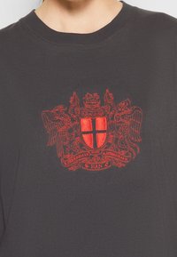 Černé bavlněné tričko s červeným vyšitým erbem se dvěma stylizovanými lvy a štítem uprostřed, doplněné zakřiveným textem.