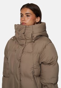 Beżowa kurtka puchowa z ociepleniem, z wysokim kołnierzem, zamykanymi na zamek kieszeniami oraz pikowanym wzorem. Wykonana z lekkiego, gładkiego materiału.