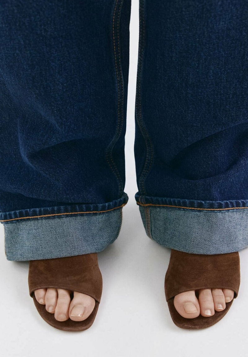 Pieds portant des sandales ouvertes en daim marron, associées à un jean denim bleu foncé retroussé, sur un fond clair.