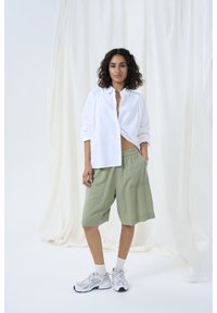 Chemise blanche à boutons, shorts verts mi-longs et baskets gris clair avec chaussettes blanches. Textures douces et coupe décontractée. Design simple.