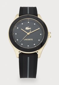 Montre avec bracelet en silicone noir et lunette dorée et noire. Le cadran rond présente des aiguilles dorées, des marqueurs en forme de diamants et un logo crocodile.