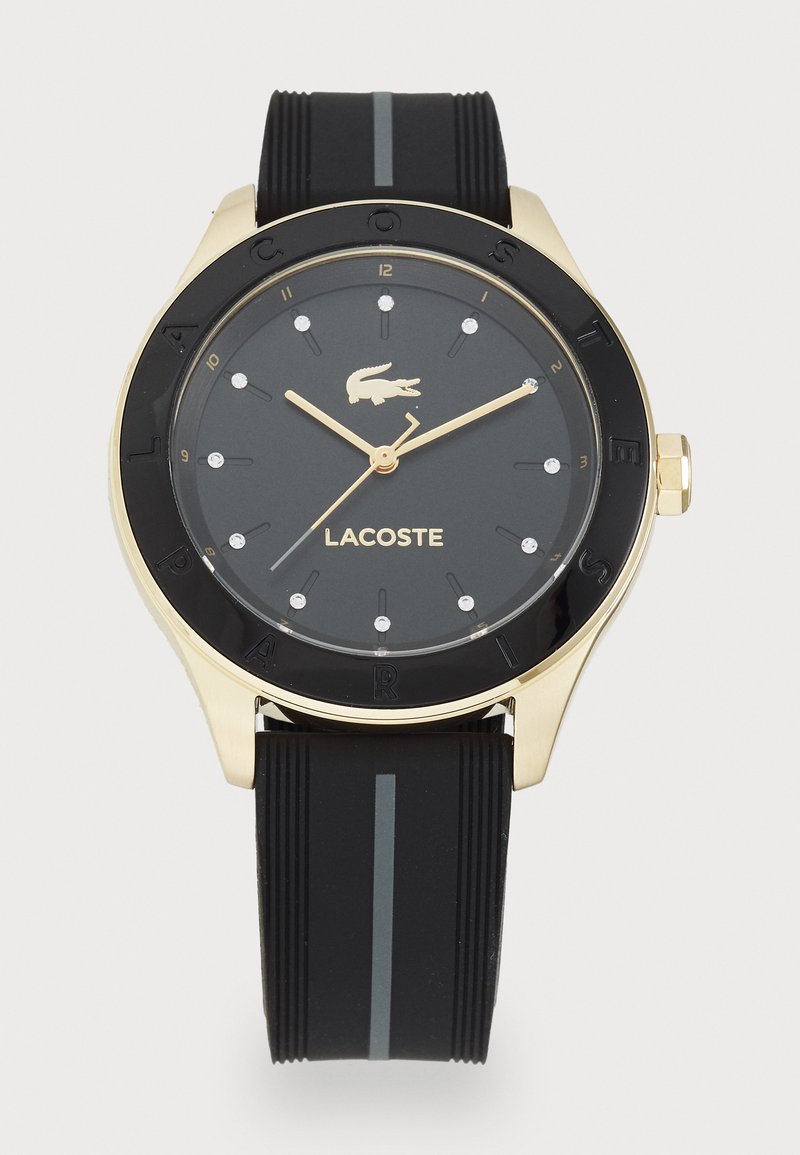 Montre avec bracelet en silicone noir et lunette dorée et noire. Le cadran rond présente des aiguilles dorées, des marqueurs en forme de diamants et un logo crocodile.