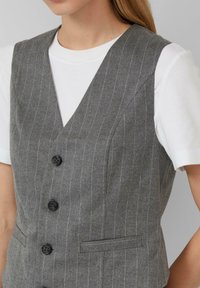 Grijze gepinde vest gemaakt van gestructureerde stof, met een diepe V-hals, zijzakken en vier zwarte knopen. Gemaakt over een wit shirt.