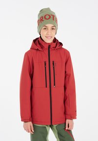 Giacca impermeabile rossa con cappuccio, due tasche con zip, accenti neri e polsini in velcro, abbinata a un berretto verde con la scritta "PROTECT".