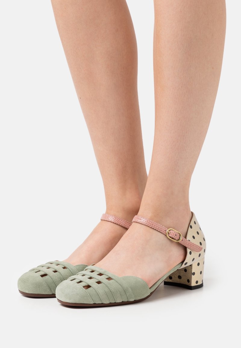 Pieds de femme portant des escarpins fermés en daim vert avec un design tissé, des brides roses à la cheville et des talons blocs beiges à pois noirs.