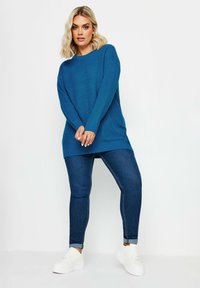 Blå stickad tröja med ribbat mönster, långa ärmar och rundad halsringning, kombinerad med mörk denimjeans och vita sneakers.