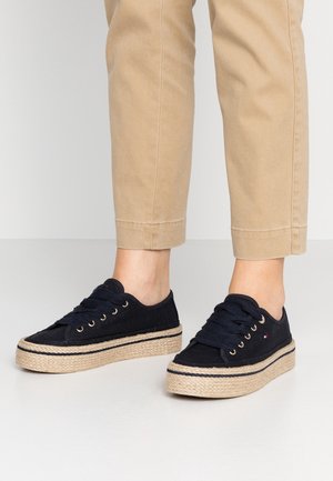 Stopy w czarnych, sznurowanych trampkach na platformie z plecioną podeszwą ze sznurka oraz beżowe spodnie o kroju skróconym, stojące na jasnym tle podłogi i ściany.