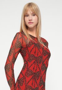 Czerwona sukienka z długimi rękawami, z czarnym wzorem w panterkę, wykonana z przezroczystego, lekkiego materiału.