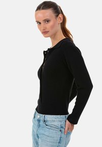 Cardigan noir en tricot à la coupe ajustée, avec un devant boutonné et un col classique. Porté avec un jean en denim bleu clair usé.