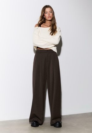 Žena s dlhými hnedými vlasmi nosí krémovo biely krátky sveter, tmavé široké nohavice a čierne platformové topánky, stojaca na neutrálnej pozadí.