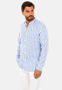 Camicia di cotone a righe blu e bianche con colletto alla coreana, maniche lunghe e chiusura con bottoni, abbinata a pantaloni bianchi.
